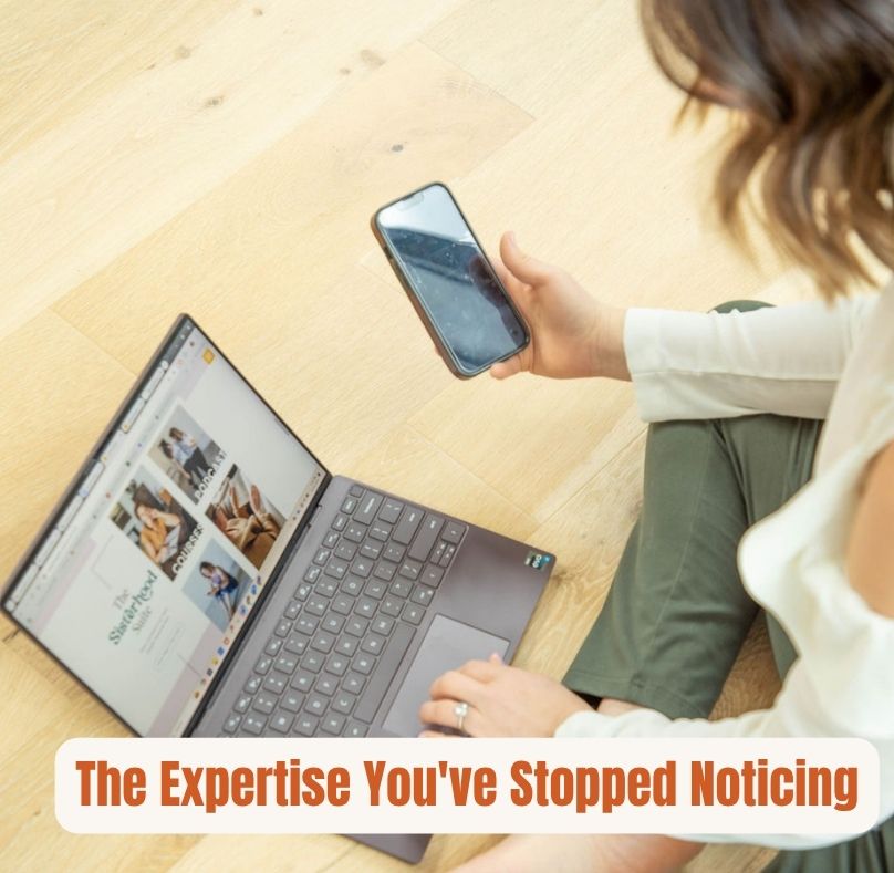 The Expertise You've Stopped Noticing — woman sitting cross-legged on a wood floor, holding a phone in one hand and typing on a laptop with the other.
